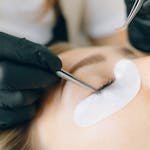 Black-gloved technician applying eyelash extensions to a woman's eye in a beauty salon setting.